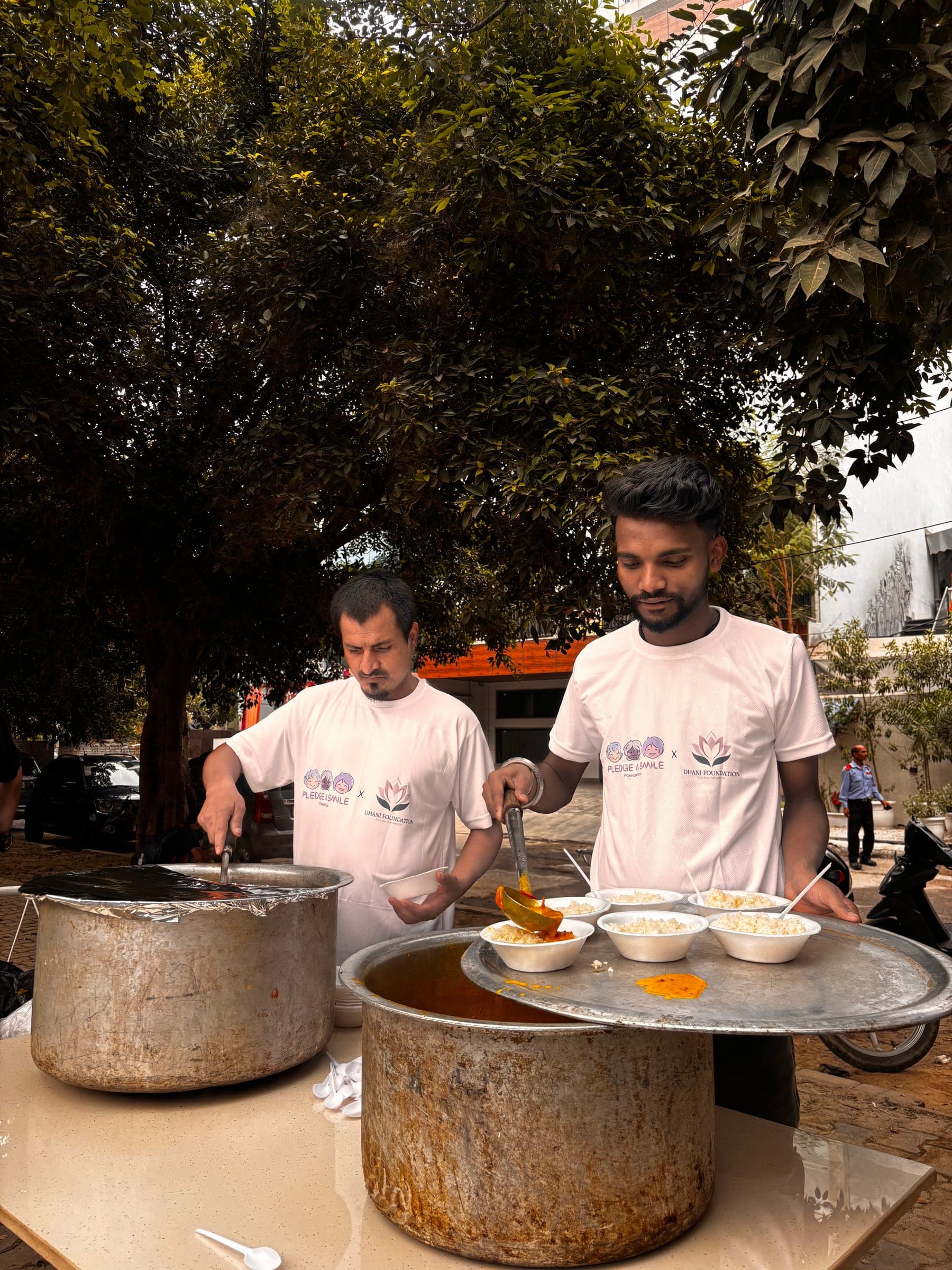 Two Dhani Foundation volunteers serve rice and curry bowls under a tree.