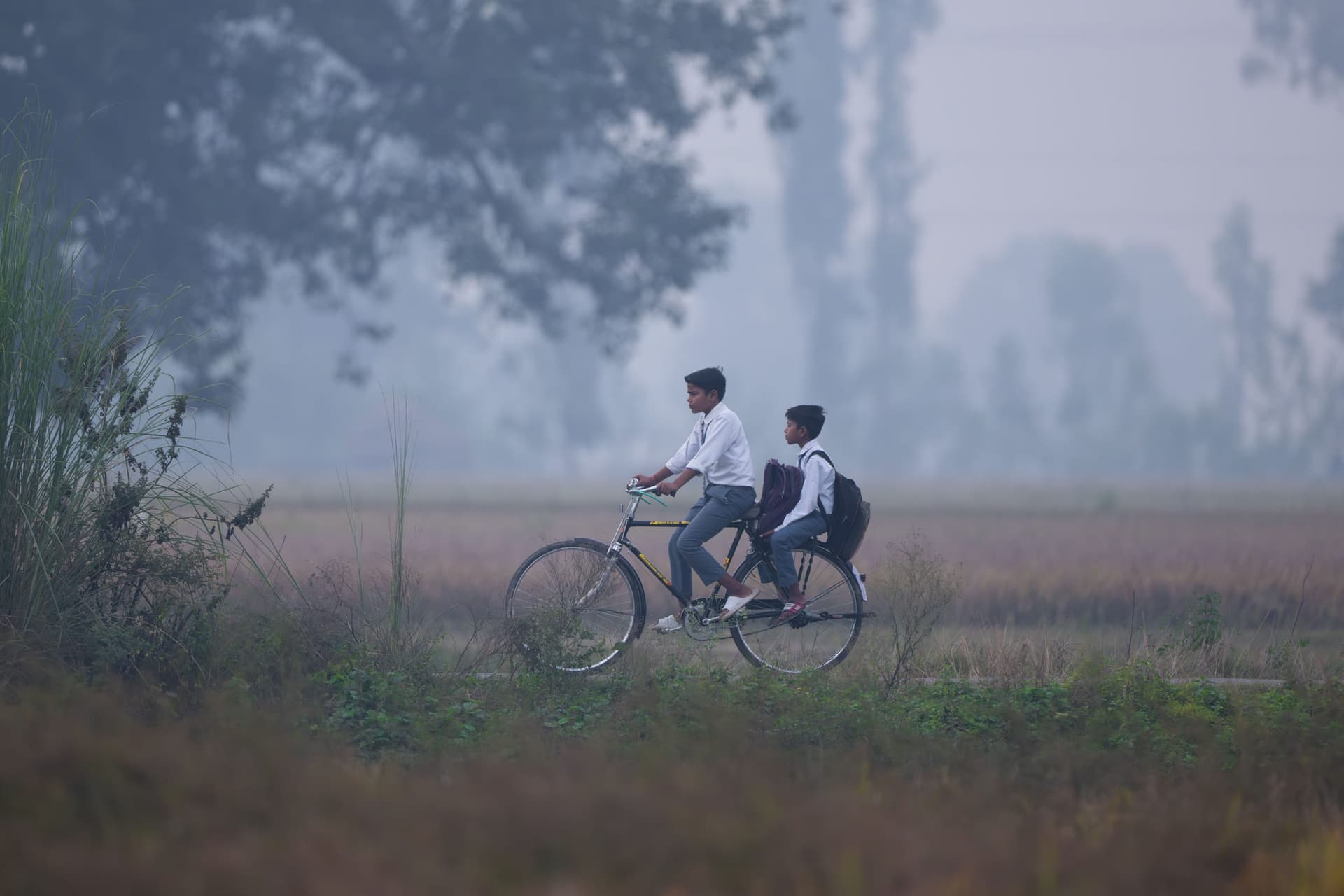 Two boys in school uniform sharing a bicycle ride through misty rural fields in early morning