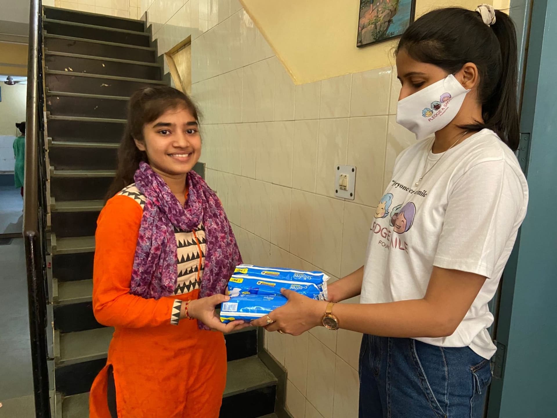 A smiling girl in an orange kurta and purple scarf receiving sanitary pads from a Pledge A Smile volunteer.