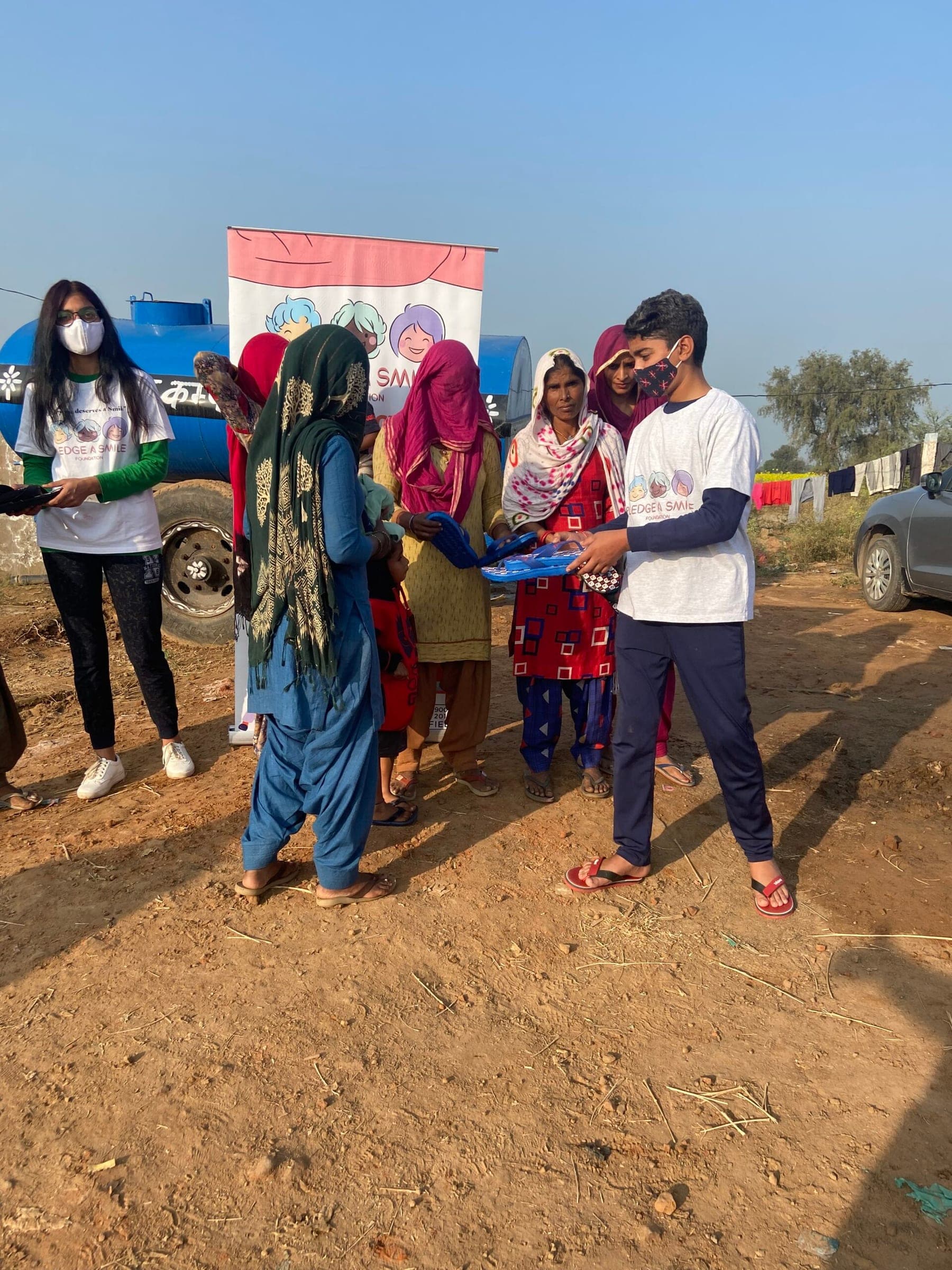 A boy hands blue sandals to a woman in a family group, the Raahat banner visible behind.
