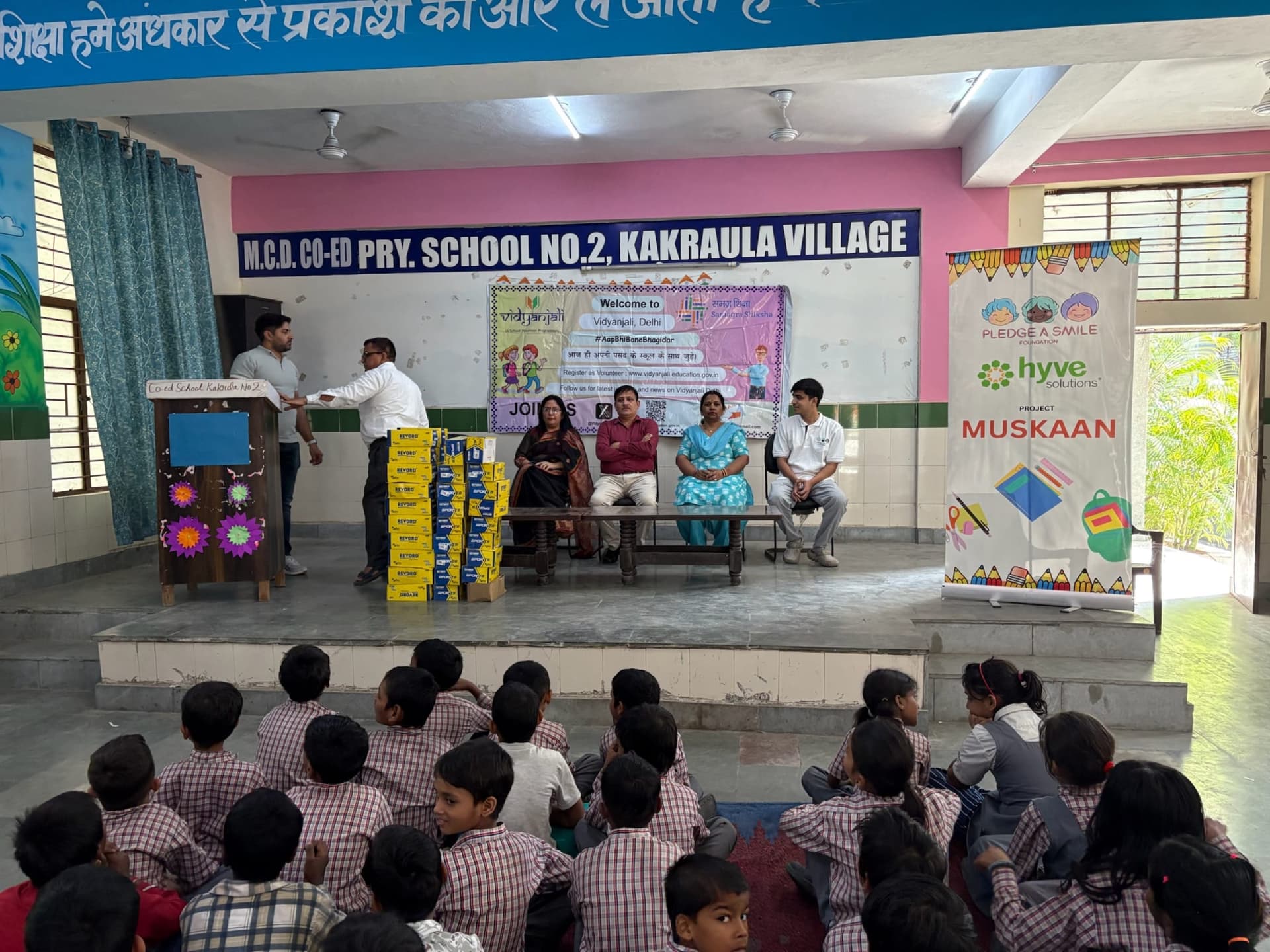 Distribution ceremony on classroom stage, kids seated watching - real mid-action moment