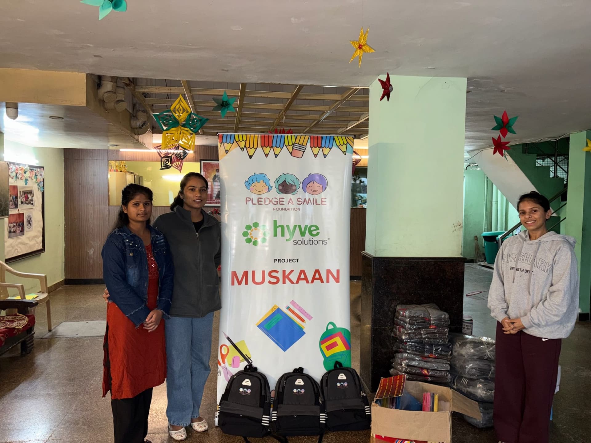 Three Pledge A Smile team members beside the Project Muskaan banner and donated backpacks