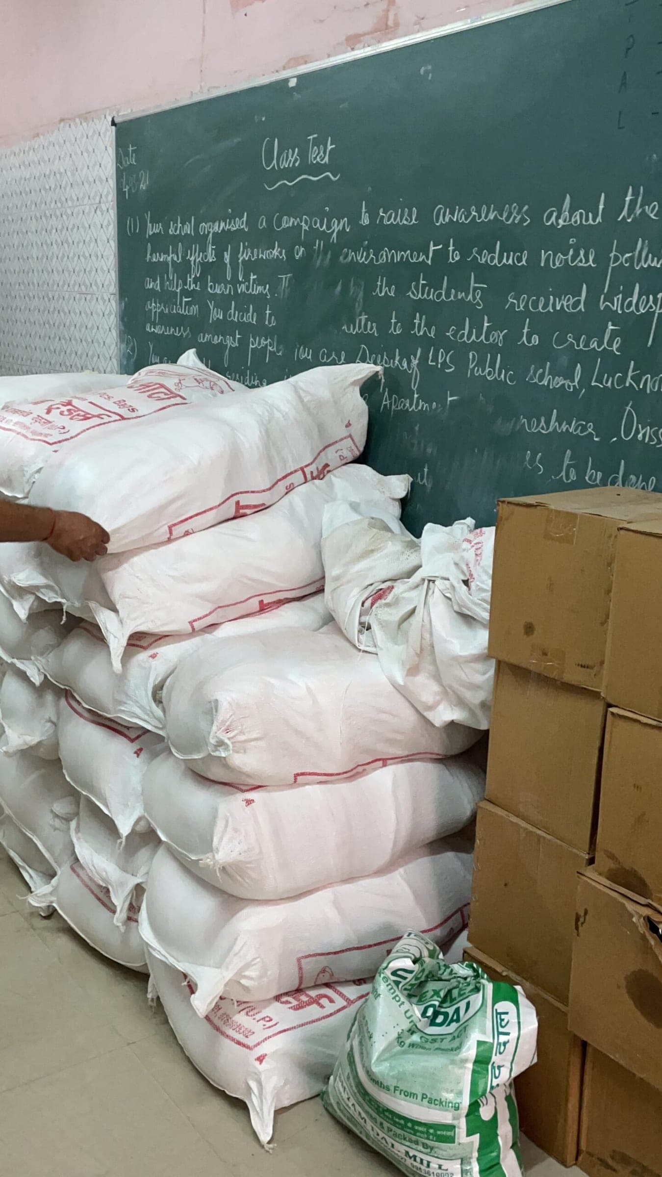 Sacks of ration grain and cartons stacked against a classroom blackboard, staged before a distribution.