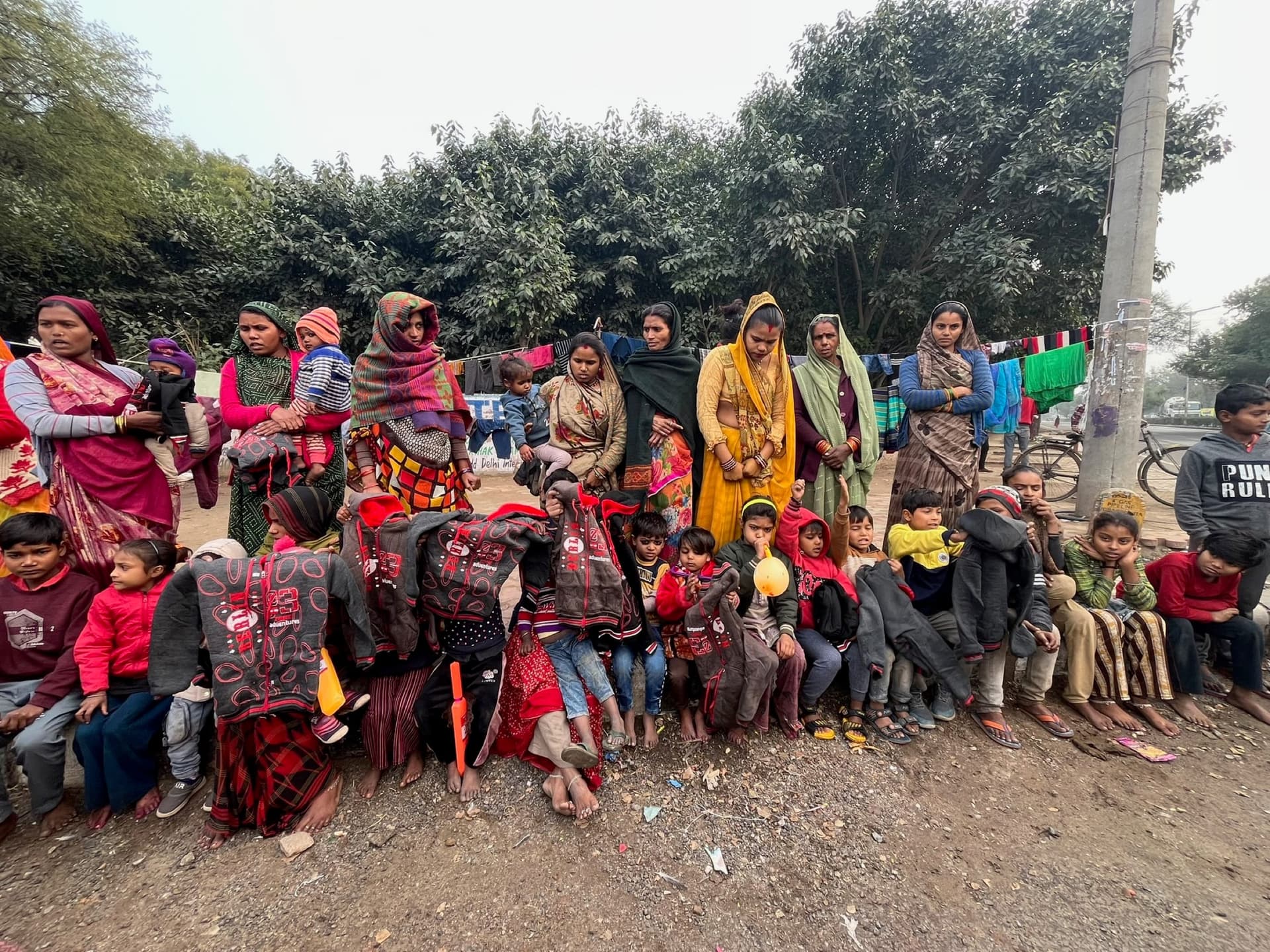Mothers and children wrapped in newly distributed grey blankets at a Delhi-NCR settlement, open winter sky across the top.