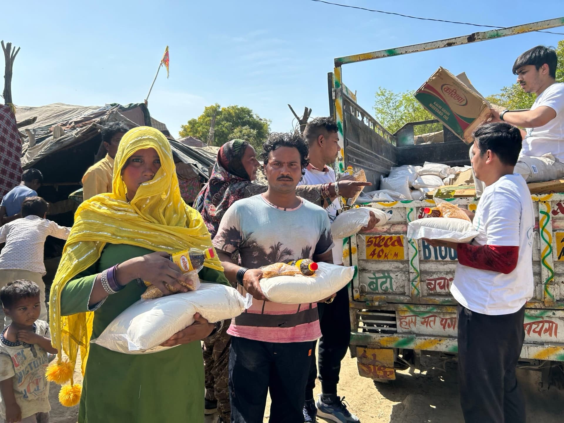 A woman in a yellow dupatta cradles her ration sack in front while volunteers unload the supply truck behind her.