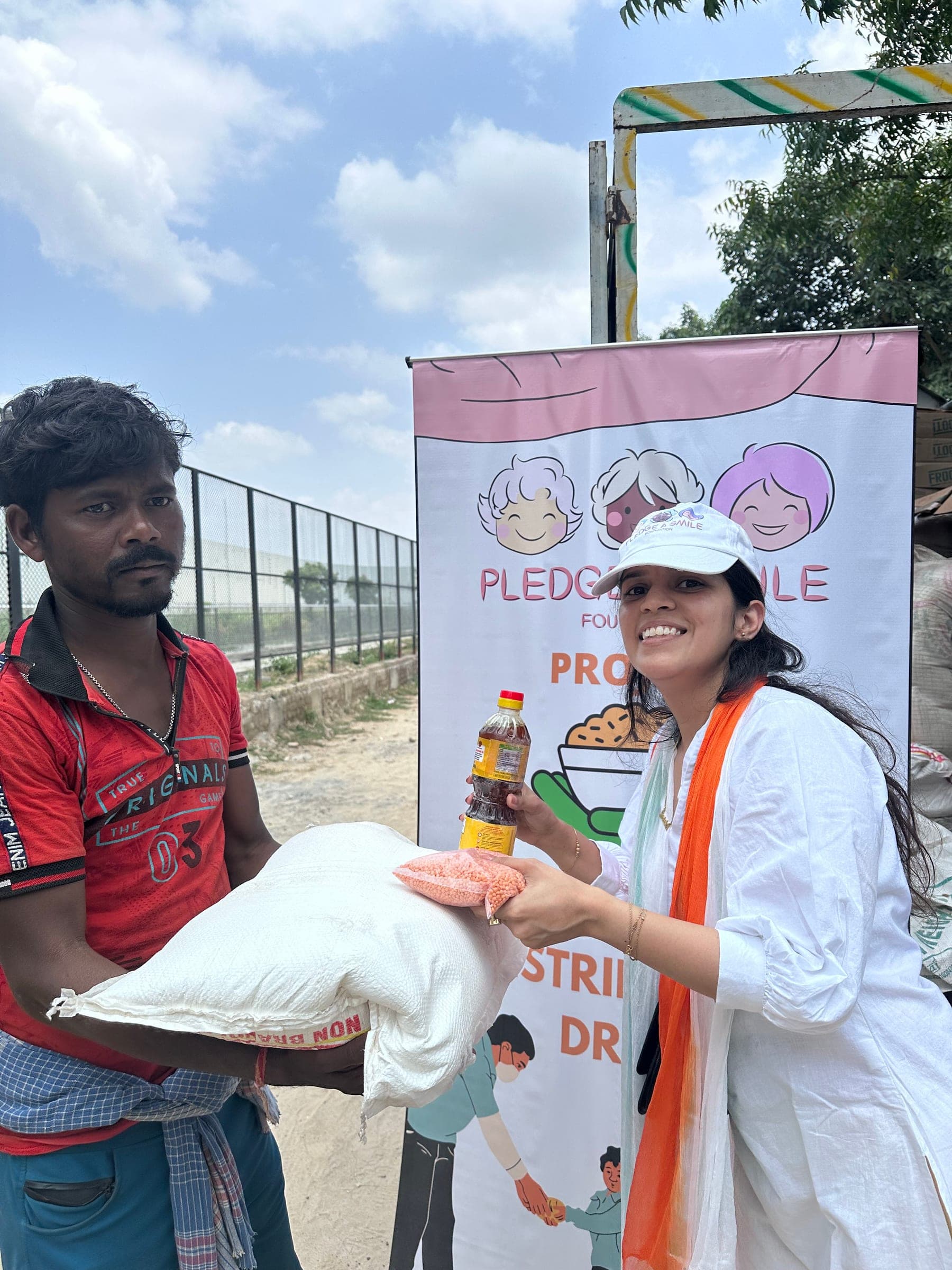 A volunteer in an orange dupatta laughs while handing a bag and bottle to a man in a red jersey.