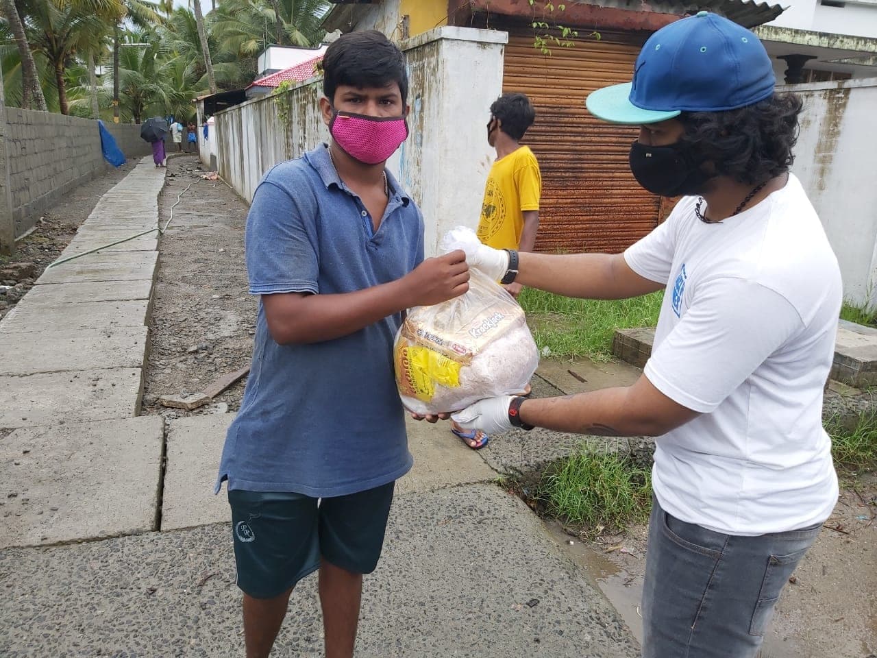 A masked teenager receives a bread-and-groceries bag from a capped volunteer at a shuttered shopfront.