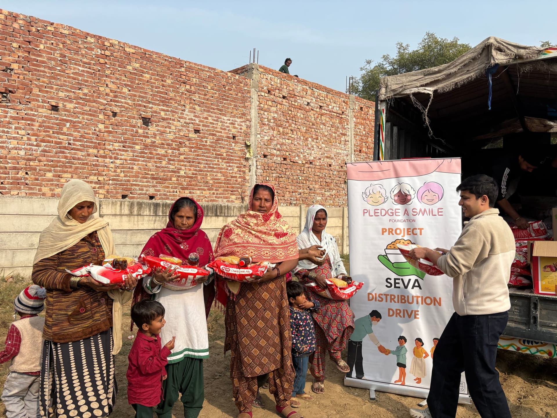 Volunteers hand atta and oil to a line of four women as two small children wait, a brick boundary wall and the Seva banner behind them.