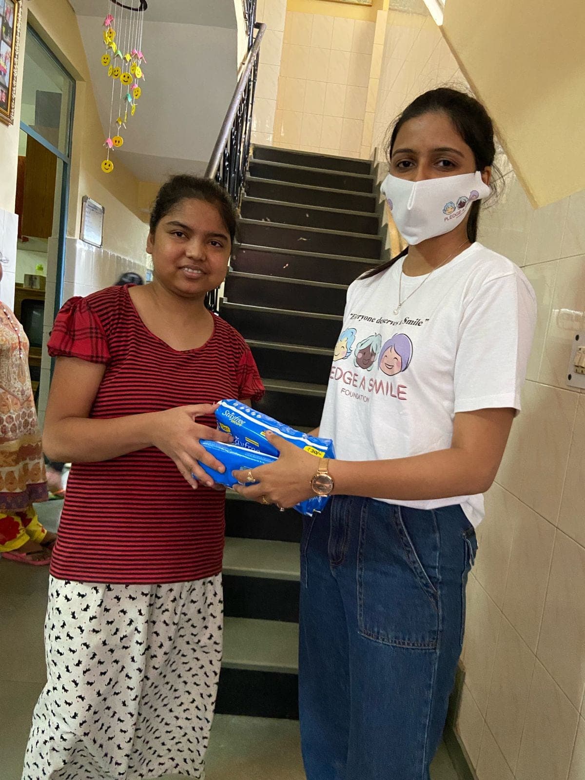 A Pledge A Smile volunteer hands sanitary-pad packets to a girl in a striped red top — the clearest branding and connection of the Noori hygiene-kit drive.