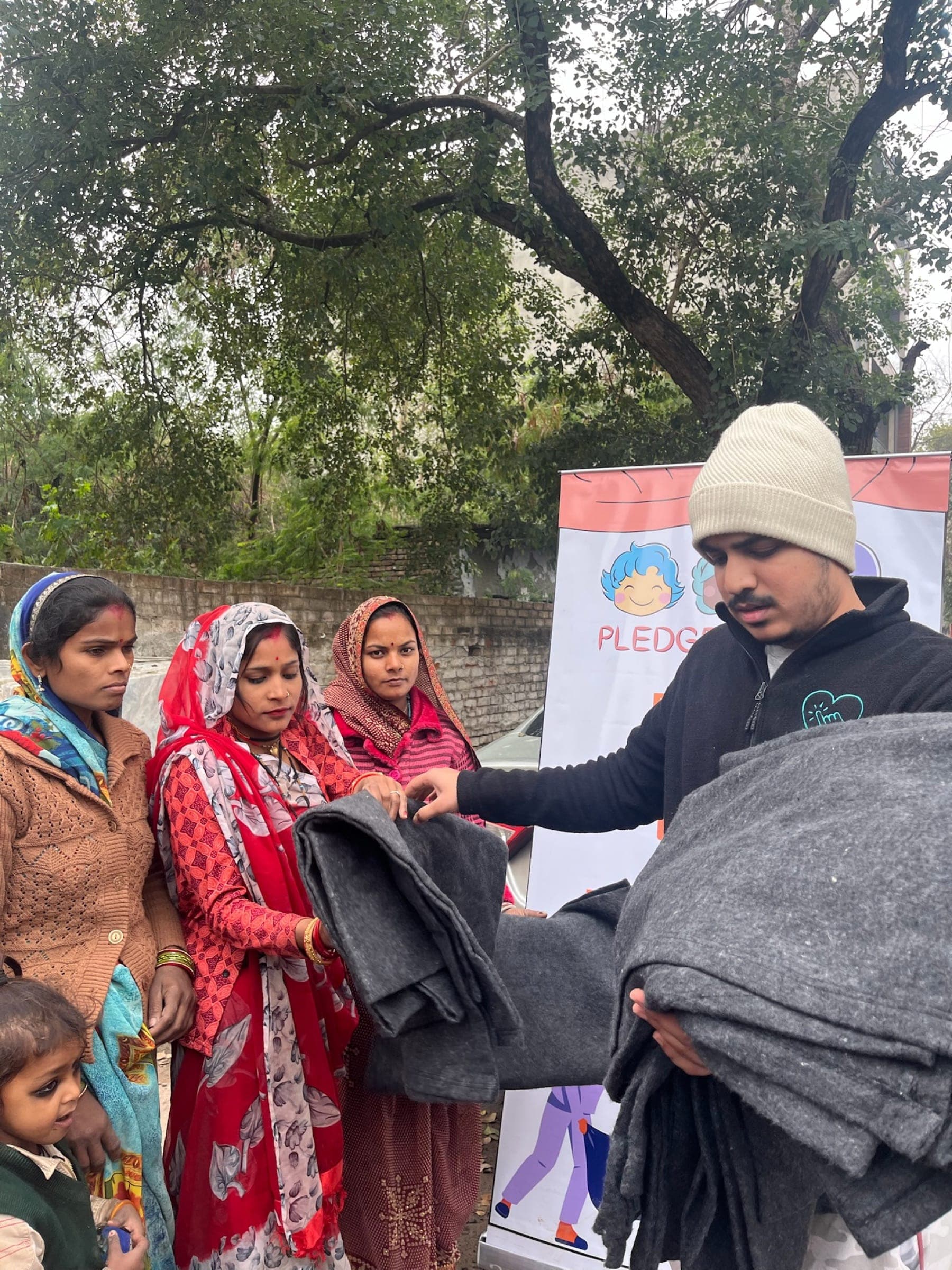 A volunteer in a beanie hands grey blankets to women in red saris at a Raahat drive