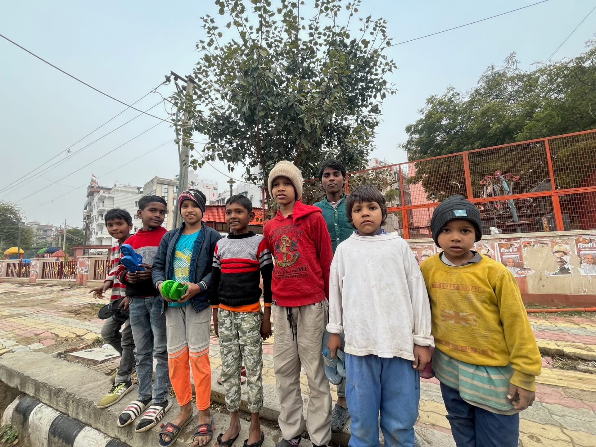 Group of boys in winter layers at a park curb, direct engaged gazes, soft hazy winter light, and a large quiet sky camera-left that comfortably holds headline text. Candid feel, no banner dominating the frame.