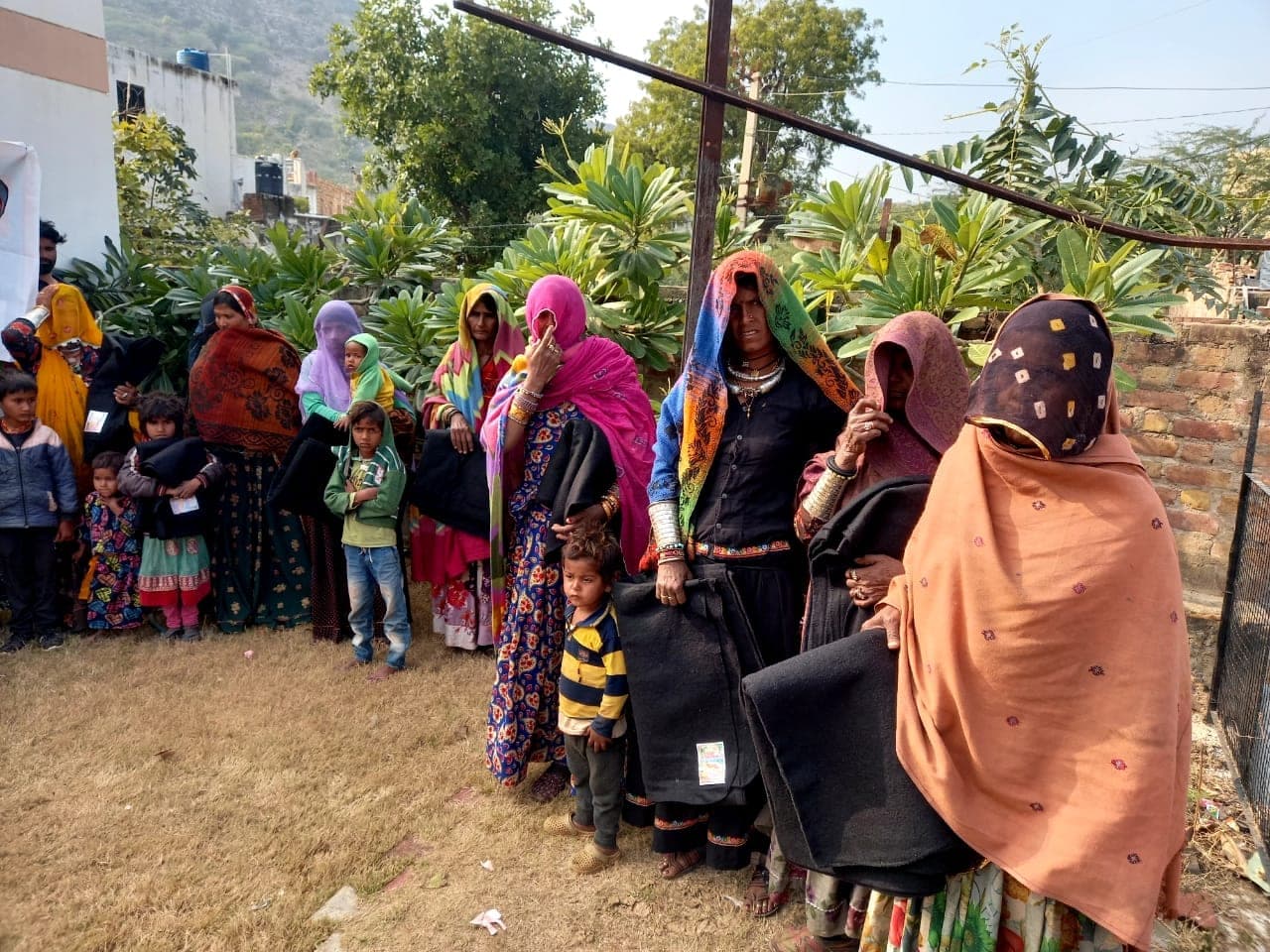 A line of about ten women in Rajasthani-style dress and silver jewellery hold folded black blankets, rocky hills behind.