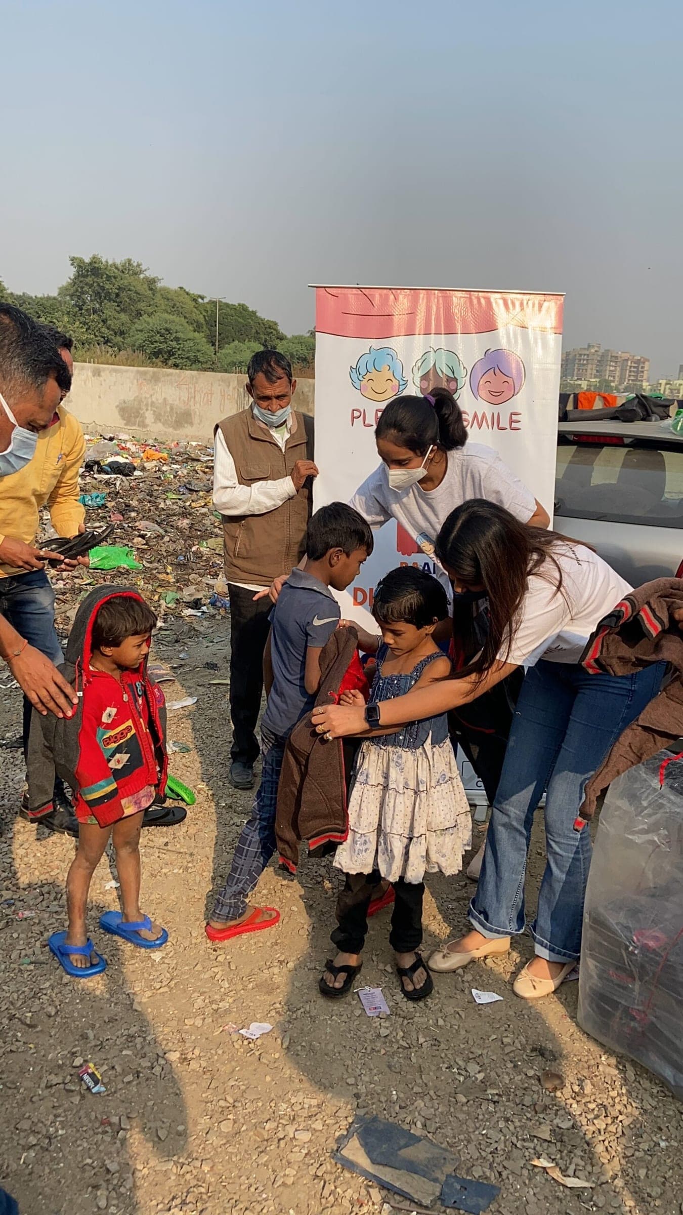 Volunteers help children into warm jackets at a landfill edge, the Project Raahat banner up, police looking on.