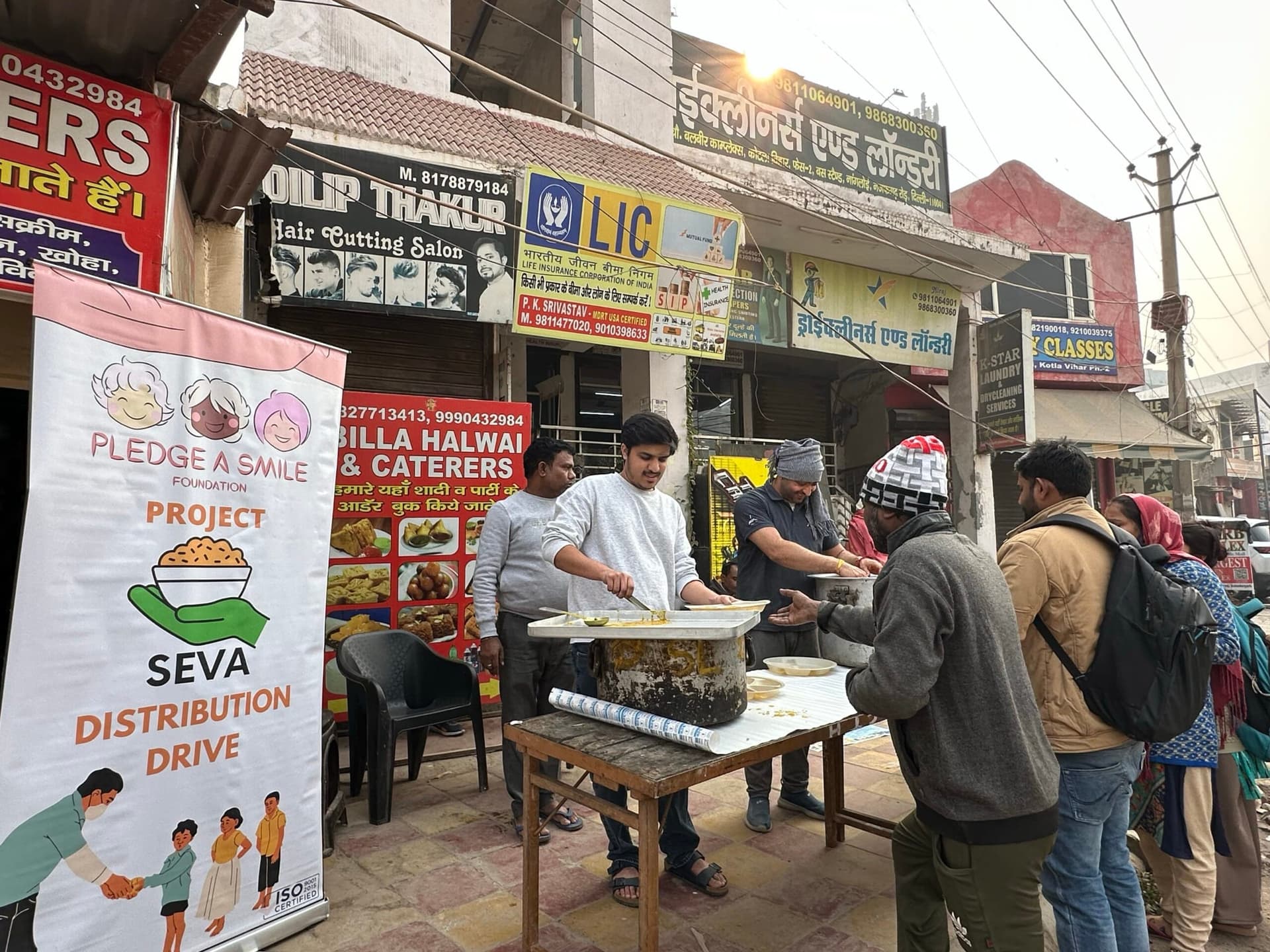 Wide scene: street-side hot food distribution table in action with SEVA standee
