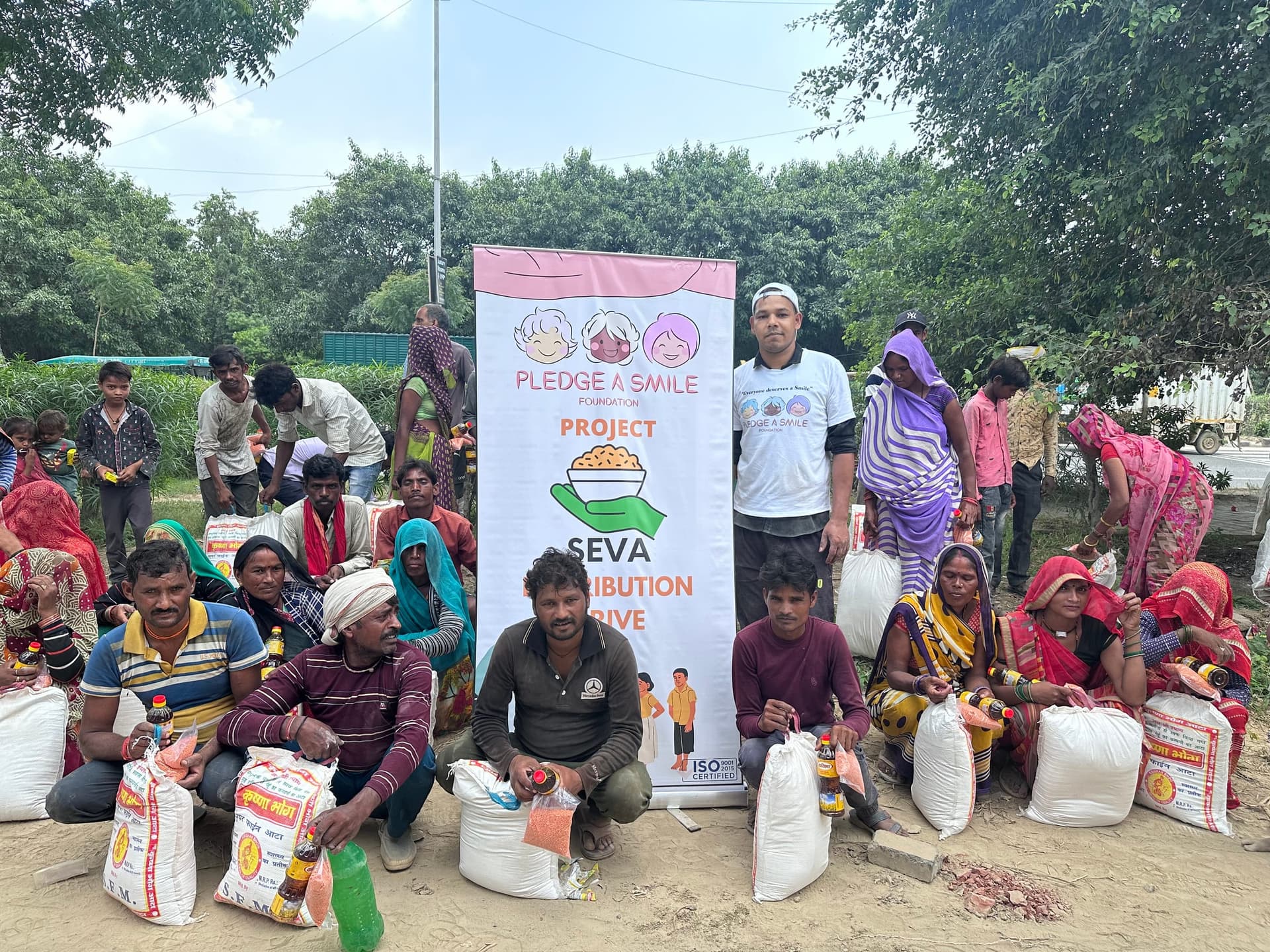 About a dozen seated recipients hold rice and oil beneath a fully legible Project Seva distribution banner, a volunteer standing at centre.
