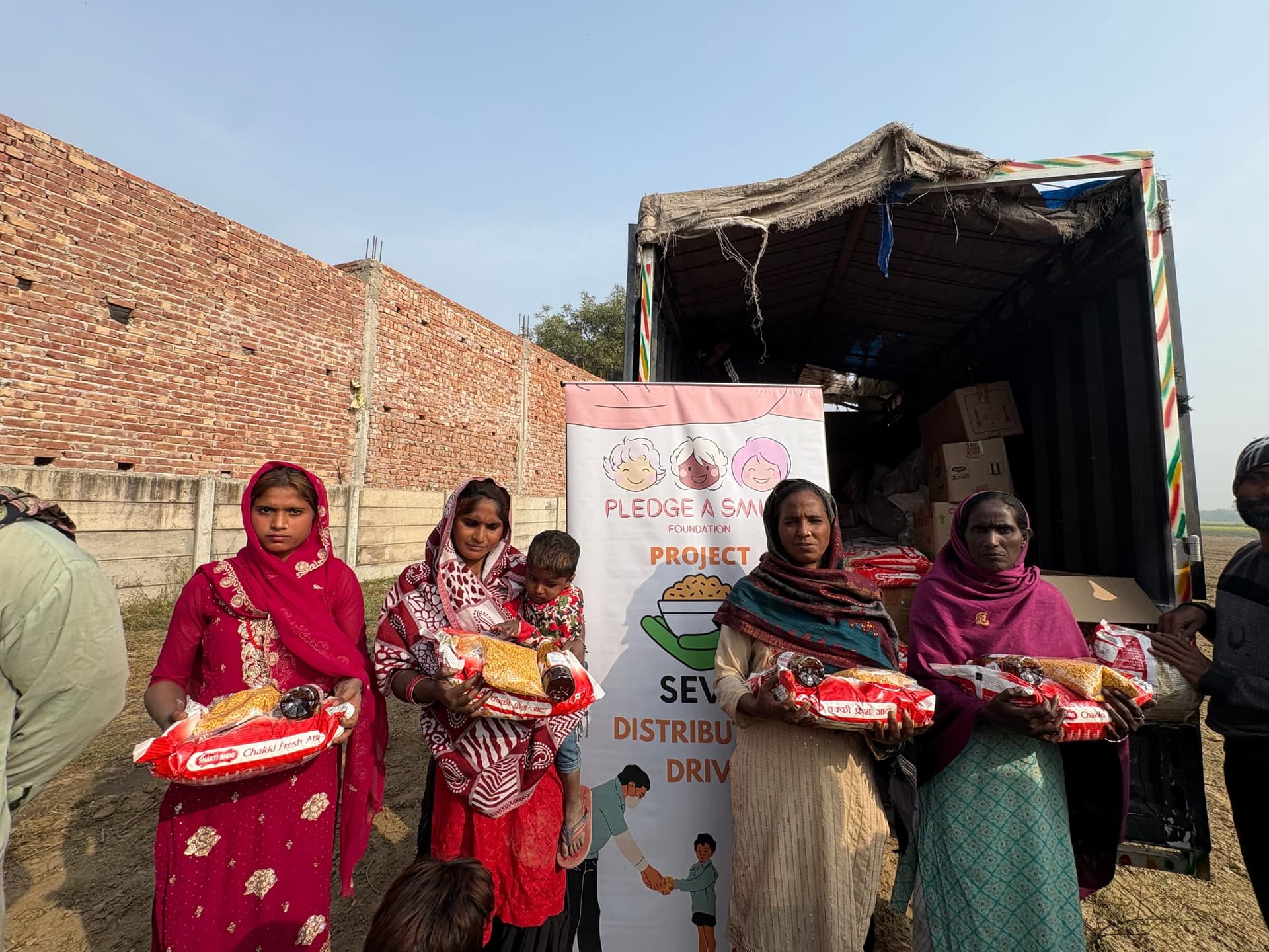 Four women in bright saris hold their atta and oil rations in a row, the Seva distribution banner and the open supply truck behind them.